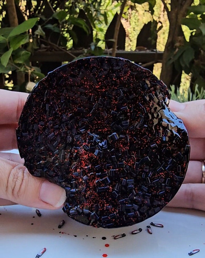 K&ouml;nnte beinhalten: Ein runder Untersetzer aus schwarzem und rotem Glitzerharz. Der Untersetzer ist mit kleinen schwarzen und roten Glitzerst&uuml;cken bedeckt.