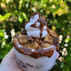 以下が含まれることがあります： 白いカップの中に、茶色のチョコレートチップワッフルが入っています。ワッフルにはホイップクリーム、チョコレートソース、チョコレートチップがトッピングされています。