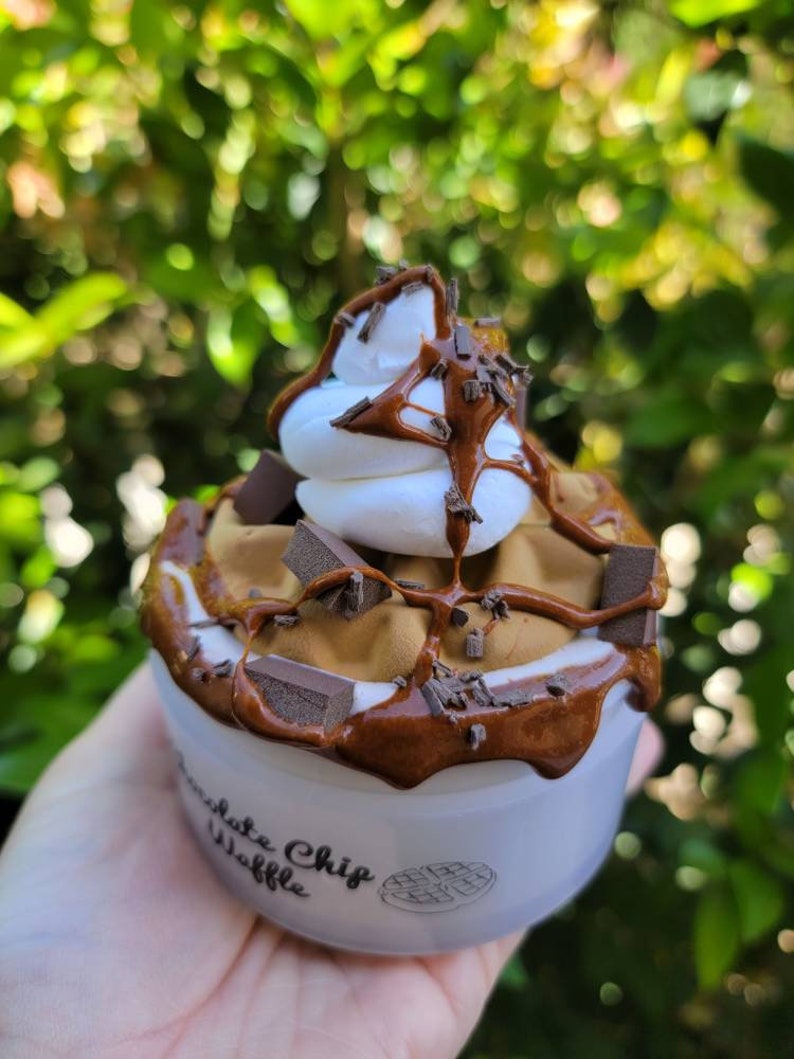 以下が含まれることがあります： 白いカップの中に、茶色のチョコレートチップワッフルが入っています。ワッフルにはホイップクリーム、チョコレートソース、チョコレートチップがトッピングされています。