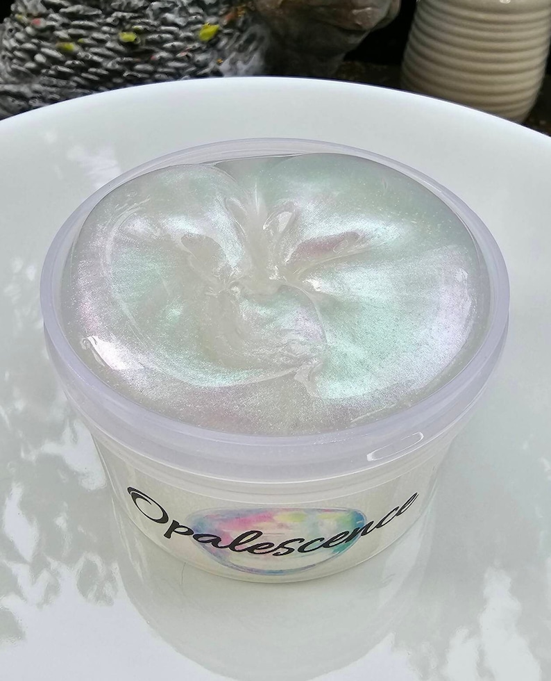 Op de afbeelding: Een doorzichtige plastic container gevuld met iriserende witte slijm. De slijm heeft een draaiende patroon en is gelabeld "Opalescence".