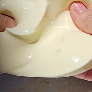 May include: A close-up of a white, creamy substance being stretched and pulled by a hand. The substance is smooth and glossy, with a slightly textured surface.