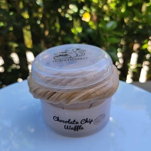 以下が含まれることがあります： 透明なプラスチック容器の中に、茶色く渦巻いたスライムが入っています。容器には、「A Crafty Monkey」と書かれた白黒のロゴが付いた蓋が付いています。容器のラベルには、「Chocolate Chip Waffle」と書かれています。