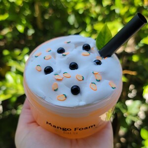 K&ouml;nnte beinhalten: Ein durchsichtiger Plastikbecher gef&uuml;llt mit einem wei&szlig;en, fluffigen Schaumtopping mit schwarzen Boba-Perlen und orangen Mangost&uuml;cken. Der Becher tr&auml;gt ein Etikett mit der Aufschrift "Mango Foam Boba Tea".