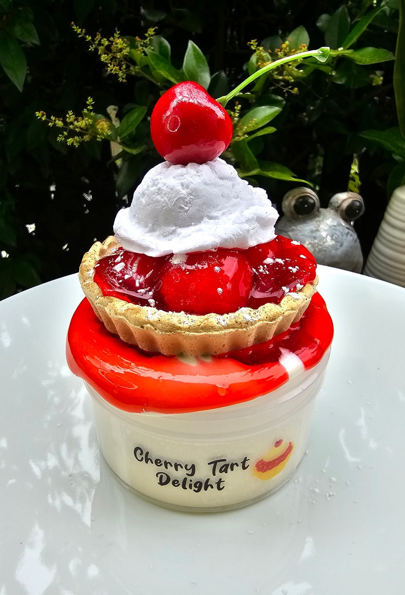 May include: A red cherry topped with white whipped cream sits on top of a cherry pie. The pie is in a white container with the words "Cherry Tart Delight" on the front.