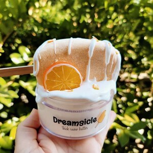 May include: A clear jar of thick snow butter with a white and orange popsicle on top. The jar has a label that says "Dreamsicle thick snow butter".