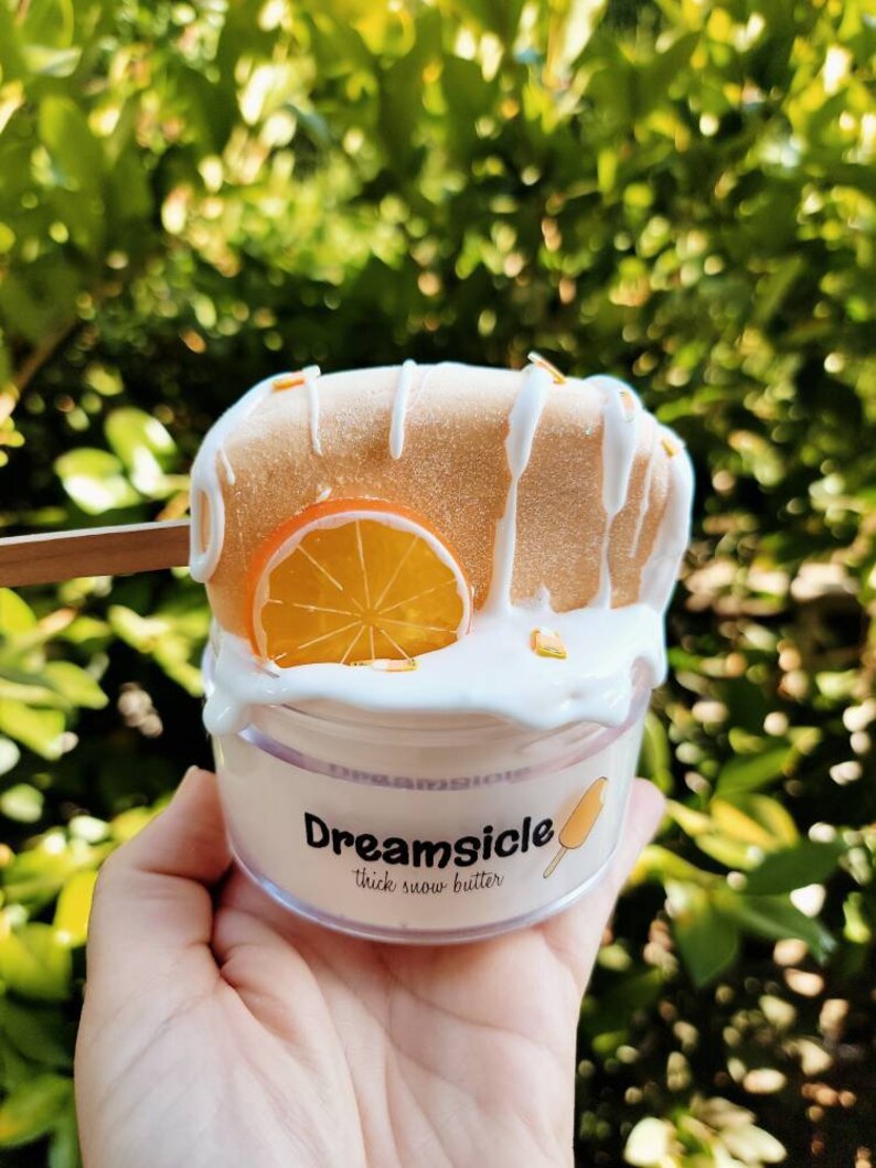 May include: A clear jar of thick snow butter with a white and orange popsicle on top. The jar has a label that says "Dreamsicle thick snow butter".