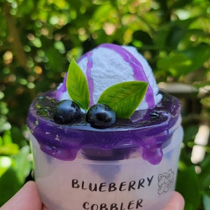 May include: A miniature blueberry cobbler dessert with a white frosting topping and purple blueberry sauce. The dessert is in a clear plastic container with the words "Blueberry Cobbler" printed on the side.