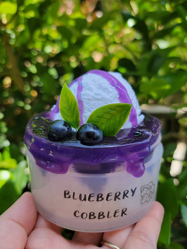 May include: A miniature blueberry cobbler dessert with a white frosting topping and purple blueberry sauce. The dessert is in a clear plastic container with the words "Blueberry Cobbler" printed on the side.