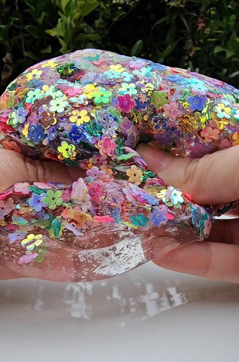May include: Clear slime with iridescent flower-shaped sequins in shades of pink, purple, blue, green, and yellow.