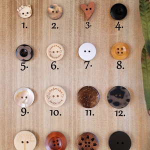 Op de afbeelding: Assortiment van 16 houten knopen in verschillende vormen, kleuren en ontwerpen. De knopen zijn gerangschikt in vier rijen van vier knopen. De bovenste rij is gelabeld "Bovenste rij aanbevolen voor dunnere banden". De knopen omvatten een kat, een hart, een cirkel, een bloem, een cirkel met de woorden "Handmade with love", een boom, een cirkel met vier gaten, een bloem en een cirkel.