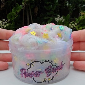 May include: A clear plastic container filled with white slime with colourful flower-shaped glitter. The container has a black and white label with the text "Flower Bomb" in purple.