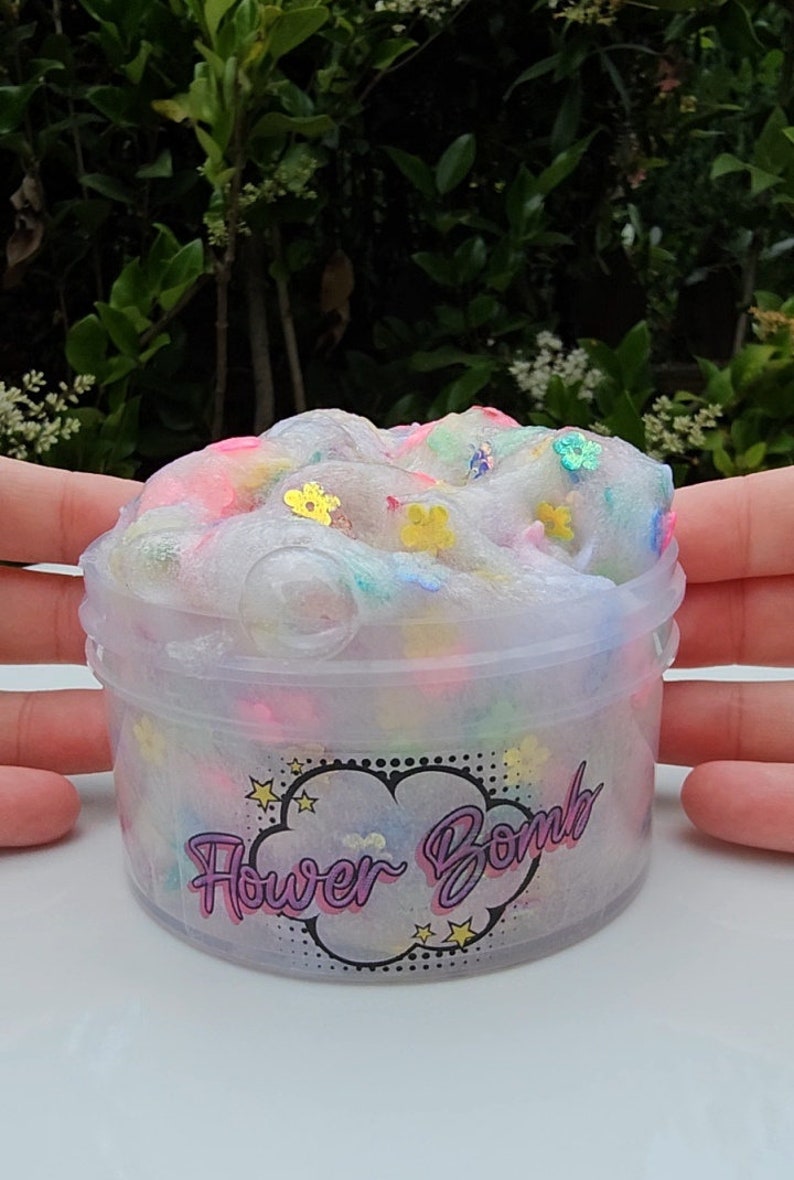 May include: A clear plastic container filled with white slime with colourful flower-shaped glitter. The container has a black and white label with the text "Flower Bomb" in purple.