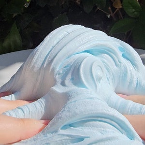 May include: A close-up of light blue slime being stretched and manipulated on a white surface. The slime has a soft, fluffy texture and is very pliable.