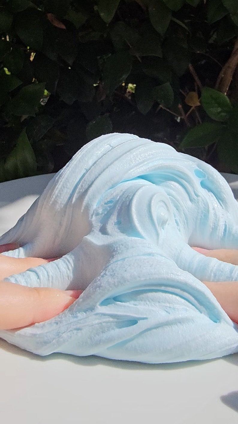 May include: A close-up of light blue slime being stretched and manipulated on a white surface. The slime has a soft, fluffy texture and is very pliable.