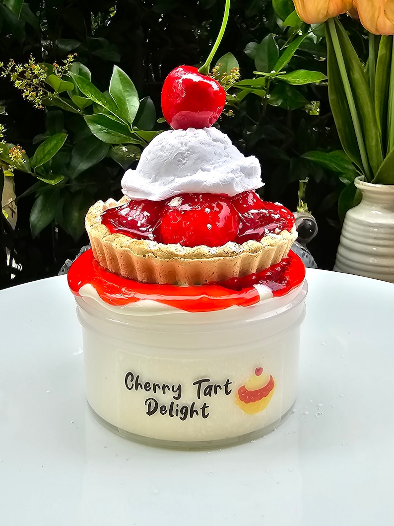 May include: A white jar of cherry tart slime with a red cherry and whipped cream topping. The jar has a label that says "Cherry Tart Delight" with a small cupcake graphic.