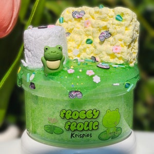 May include: Green slime with a frog charm, a white marshmallow, and a yellow foam with butterflies and flowers. The slime jar has a green label that says "Froggie Frolic Krispies".