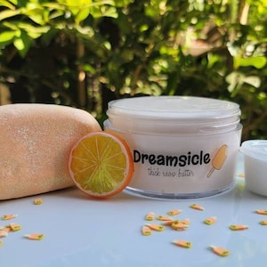 May include: A white jar of thick snow butter with the label "Dreamsicle" and a small orange slice. The jar is sitting on a white surface with orange and yellow sprinkles.