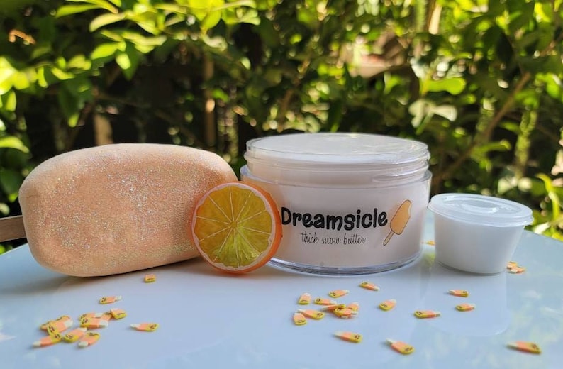 May include: A white jar of thick snow butter with the label "Dreamsicle" and a small orange slice. The jar is sitting on a white surface with orange and yellow sprinkles.