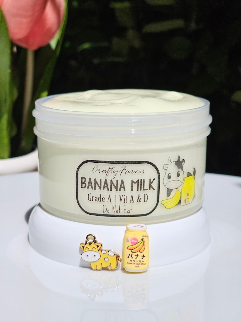 May include: A white jar with a yellow lid containing a light yellow banana milk product. The jar has a label that reads "Crafty Farms Banana Milk Grade A | Vit A & D Do Not Eat". There is a small yellow cow charm and a miniature yellow banana milk carton with Japanese text.
