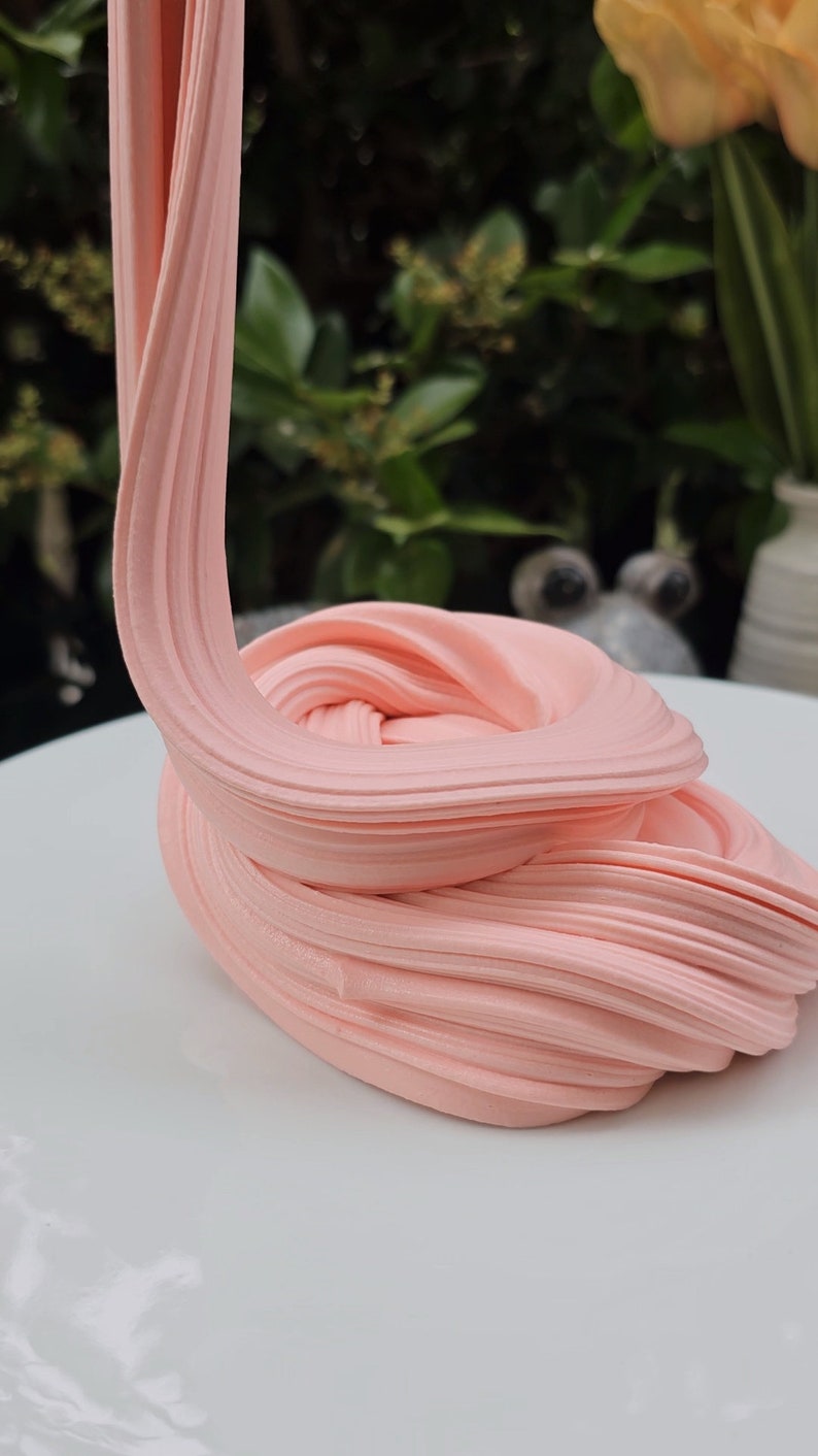 May include: A close-up of pink slime being stretched and pulled on a white surface. The slime is soft and smooth, and it has a slightly glossy finish.