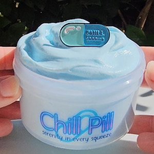 May include: A clear plastic container of light blue slime with a blue and green pill-shaped pin on top. The container has the text "Chill Pill" in a blue and purple font with the words "serenity in every squeeze" underneath.