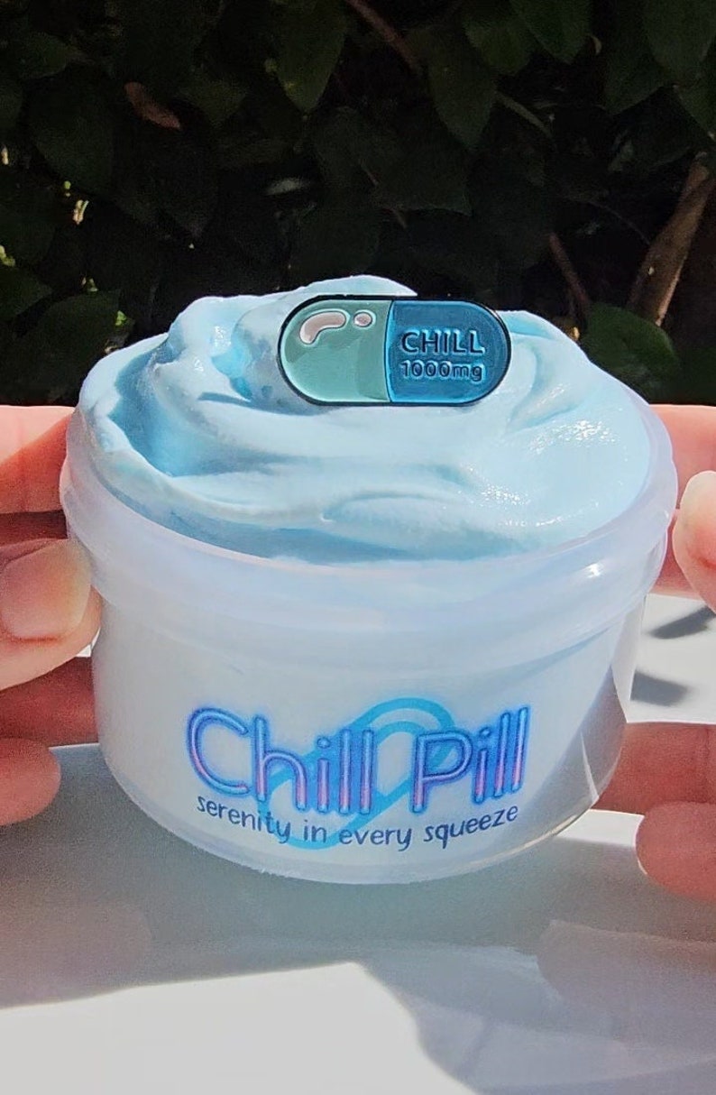 May include: A clear plastic container of light blue slime with a blue and green pill-shaped pin on top. The container has the text "Chill Pill" in a blue and purple font with the words "serenity in every squeeze" underneath.