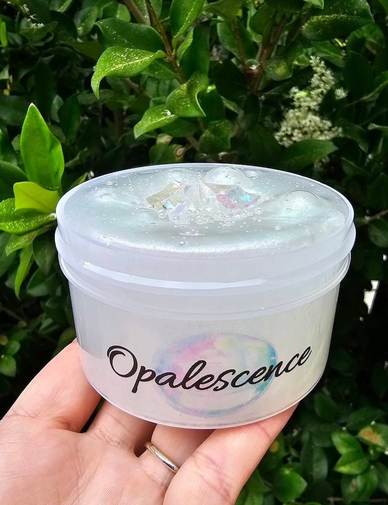 Op de afbeelding: Een doorzichtige plastic container gevuld met witte slijm met iriserende glitter en een grote iriserende kristal. De container heeft een zwart label met het woord "Opalescence" geschreven in een cursief schrift.