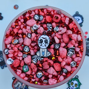 May include: A clear glass jar filled with red slime and various horror movie character charms. The charms include a masked killer, a clown, and a ghost. The jar also contains red and silver glitter.