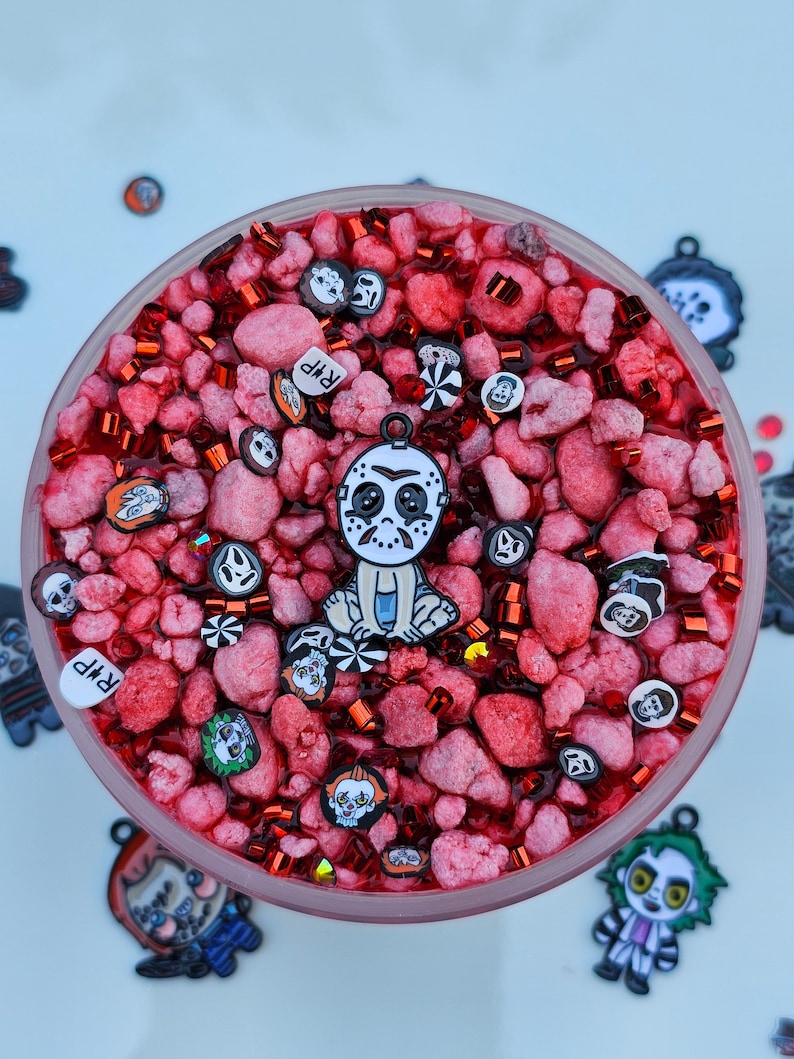 May include: A clear glass jar filled with red slime and various horror movie character charms. The charms include a masked killer, a clown, and a ghost. The jar also contains red and silver glitter.