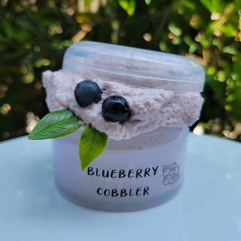 May include: A clear plastic container filled with a purple blueberry cobbler dessert. The container has a white label with the text "Blueberry Cobbler" printed on it.