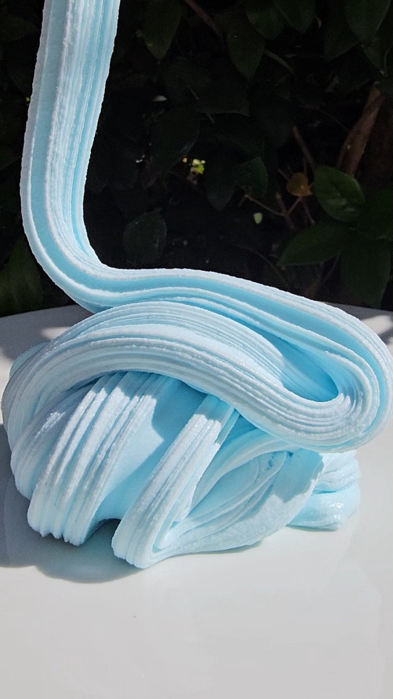 May include: A close-up of light blue slime, a popular children's toy, on a white surface. The slime is soft and stretchy, and it has a smooth, glossy finish.