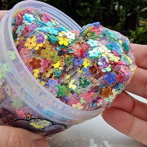 May include: A clear slime with iridescent flower-shaped glitter inside a plastic container. The slime is being held by a hand and is being pulled out of the container.