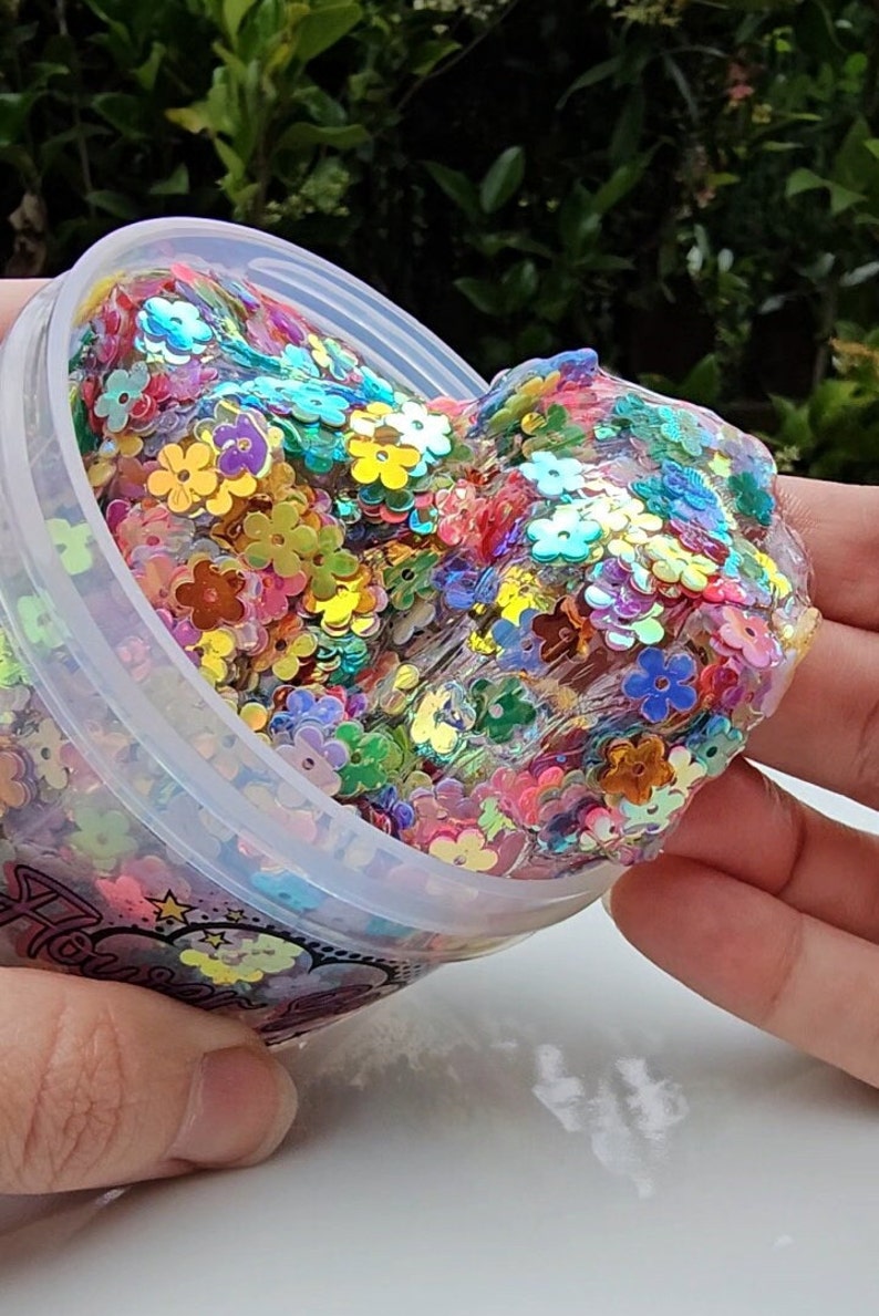 May include: A clear slime with iridescent flower-shaped glitter inside a plastic container. The slime is being held by a hand and is being pulled out of the container.