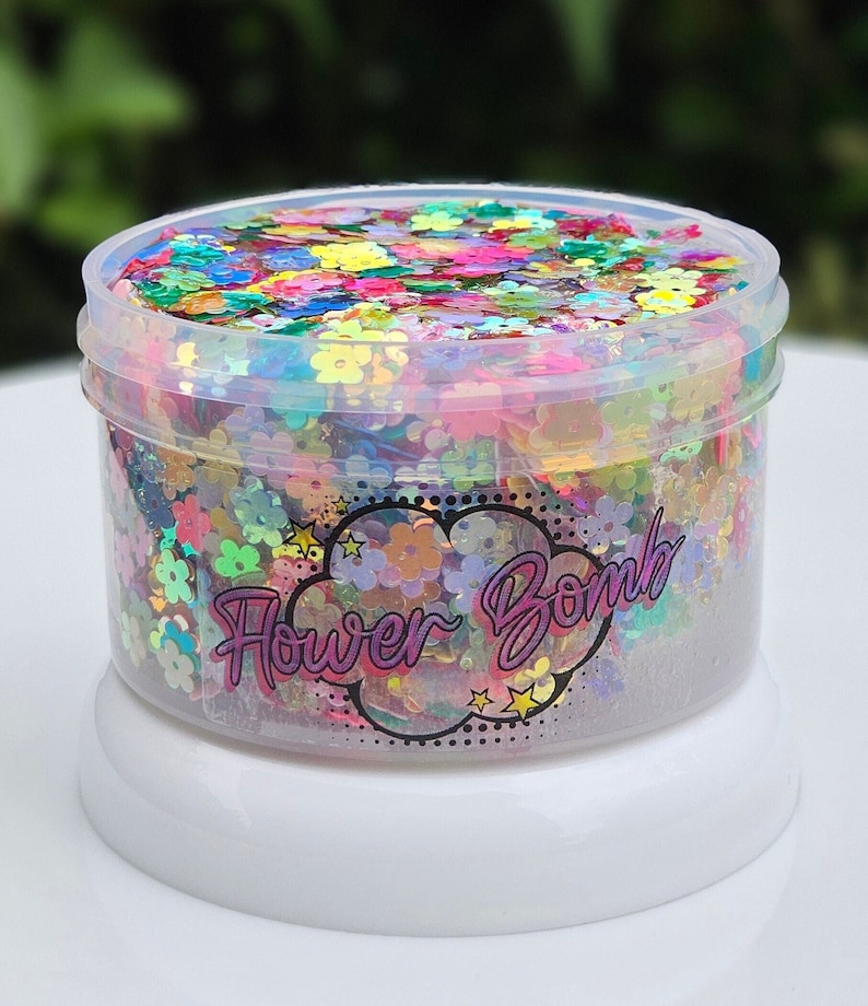 May include: A clear plastic container filled with colourful iridescent flower-shaped glitter. The container has a white lid and a label that reads "Flower Bomb".