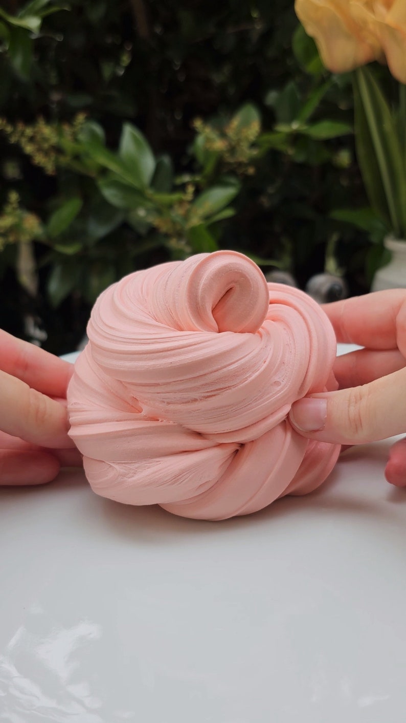 May include: A close-up of a pink slime being stretched and manipulated by hands. The slime is soft and smooth, and it has a light pink color.