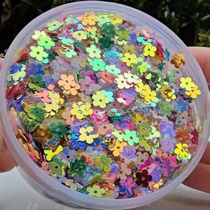 May include: A clear plastic container filled with iridescent flower-shaped sequins in various colours, including pink, green, yellow, and blue.