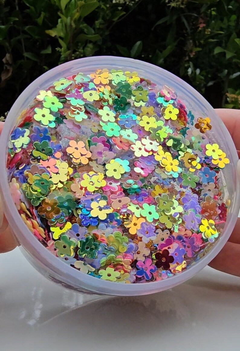 May include: A clear plastic container filled with iridescent flower-shaped sequins in various colours, including pink, green, yellow, and blue.