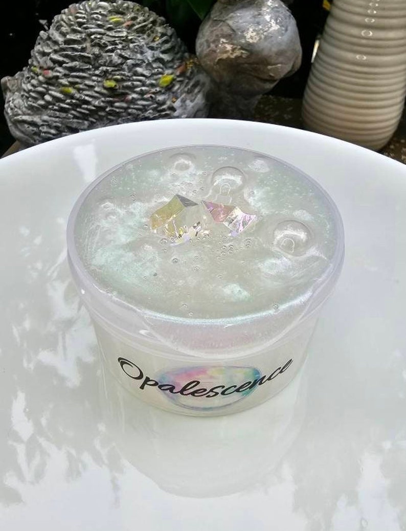 Op de afbeelding: Een heldere slijm met iriserende glitter en kleine, doorzichtige plastic vormen in een witte plastic container met een zwart label waarop "Opalescence" staat.