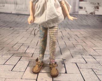 Op de afbeelding: Een pop met een witte jurk en bruine schoenen. De benen van de pop zijn bedekt met leggings met patronen met tekst en bloemmotieven. De schoenen hebben een band- en gespsluiting.