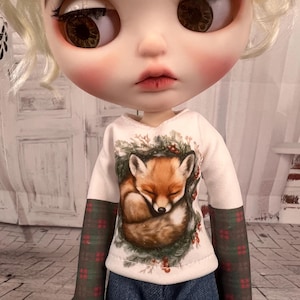 May include: A doll wearing a white long-sleeved shirt with a sleeping fox graphic. The sleeves are patterned with a green and red plaid design. The doll is wearing blue jeans.