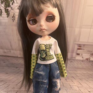 Op de afbeelding: Een pop met lang grijs haar en grote ogen, gekleed in een wit shirt met de tekst "LET ME BE FRANK" en een afbeelding van Frankenstein's monster. De pop draagt ook een blauwe jeans met distressed details.