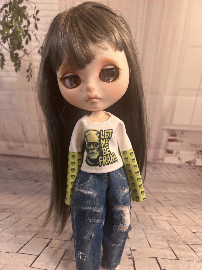 Op de afbeelding: Een pop met lang grijs haar en grote ogen, gekleed in een wit shirt met de tekst "LET ME BE FRANK" en een afbeelding van Frankenstein's monster. De pop draagt ook een blauwe jeans met distressed details.