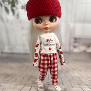 Op de afbeelding: Een pop met een rode gehaakte hoed met een groene pompon, een wit shirt met de tekst "Blythe Pick Your Own Apple Orchard" en een rood-witte geruite broek. De pop draagt witte sneakers.