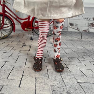 May include: A doll wearing a white dress with red and white striped knee-high socks on one leg and white socks with a ladybug pattern on the other leg. The doll is wearing brown shoes.