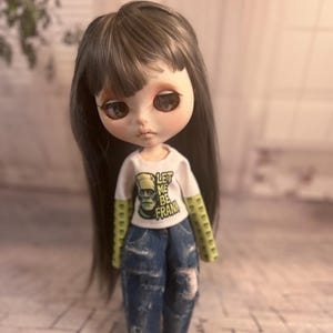 Op de afbeelding: Een pop met lang, donker haar en grote ogen, gekleed in een wit t-shirt met de tekst "LET ME BE FRANK" en een afbeelding van Frankenstein. De pop draagt ook een blauwe jeans met distressed details.