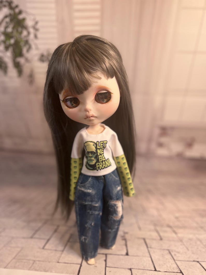 Op de afbeelding: Een pop met lang, donker haar en grote ogen, gekleed in een wit t-shirt met de tekst "LET ME BE FRANK" en een afbeelding van Frankenstein. De pop draagt ook een blauwe jeans met distressed details.
