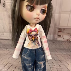 May include: A doll with long gray hair and large eyes wearing a white long-sleeved shirt with a colorful owl graphic and pink plaid sleeves. The doll is also wearing blue denim jeans with distressed details.