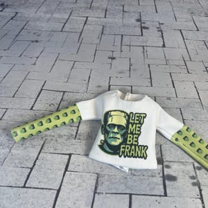 Op de afbeelding: Wit shirt met lange mouwen met een afbeelding van Frankenstein en de tekst "LET ME BE FRANK". De mouwen zijn lichtgroen met een herhalend vierkant patroon.