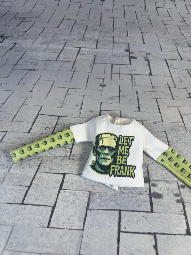 Op de afbeelding: Wit shirt met lange mouwen met een afbeelding van Frankenstein en de tekst "LET ME BE FRANK". De mouwen zijn lichtgroen met een herhalend vierkant patroon.