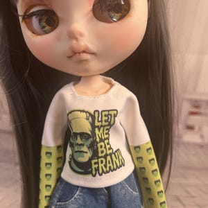 Op de afbeelding: Een pop met lang grijs haar en grote ogen draagt een wit shirt met lange mouwen met de tekst "LET ME BE FRANK" en een afbeelding van Frankenstein's monster. De mouwen zijn groen met een herhalend Frankenstein's monster ontwerp. De pop draagt ook een spijkerbroek short.